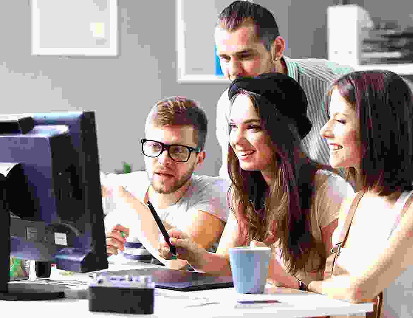 Quatre jeunes collègues, deux hommes et deux femmes, se rassemblent autour d'un écran d'ordinateur en souriant, avec une tasse bleue et une tablette graphique sur le bureau.