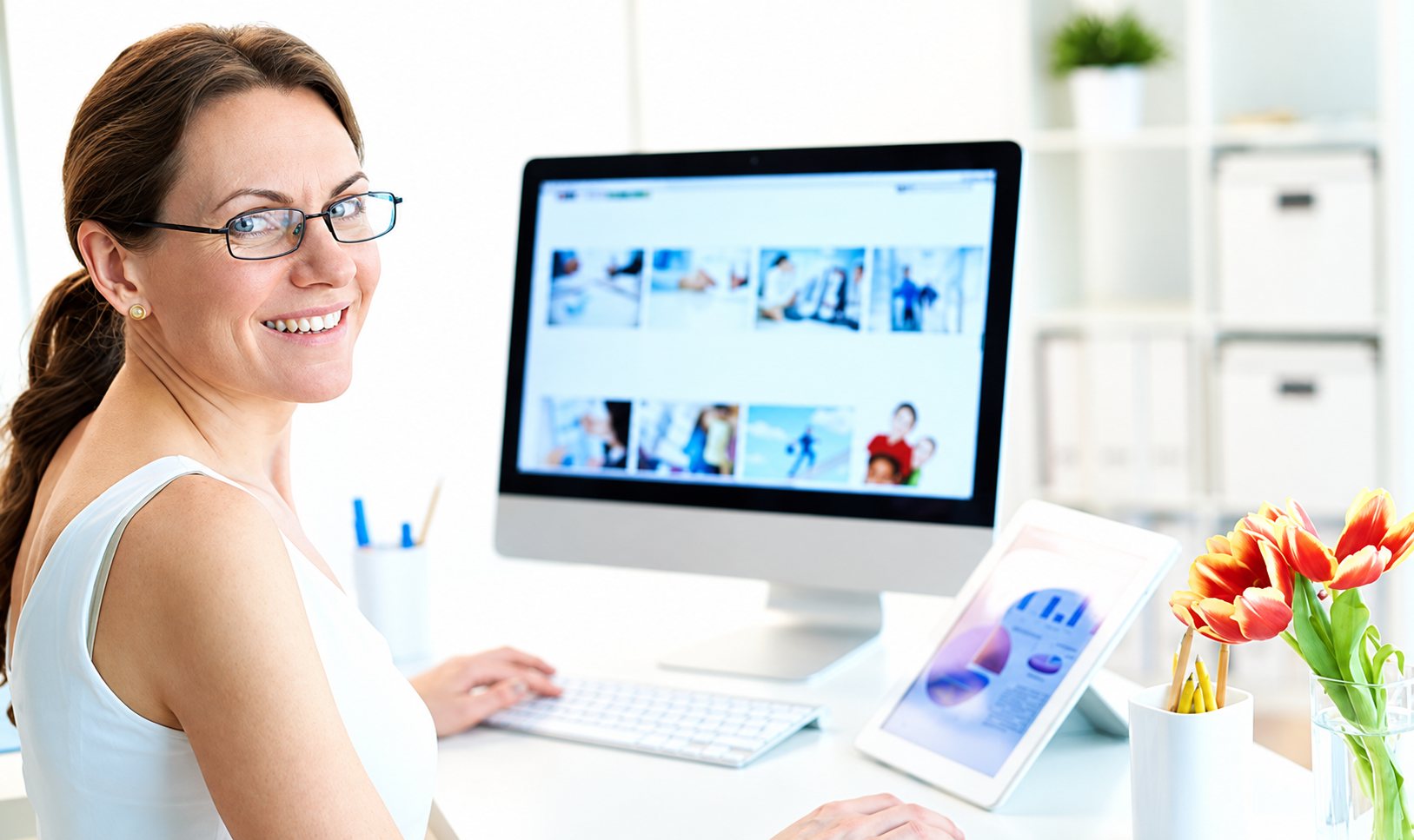 Une femme souriante portant des lunettes est assise à un bureau lumineux devant un grand écran d'ordinateur, avec une tablette et un vase de tulipes à côté.