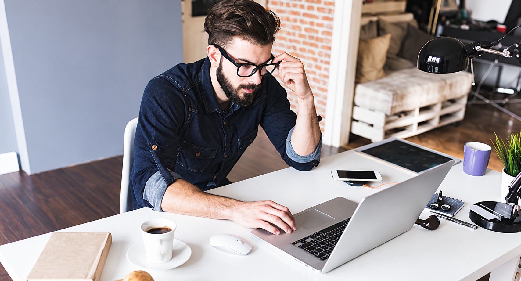 Un homme barbu portant des lunettes et une chemise sombre travaille sur un ordinateur portable posé sur un bureau blanc, avec une tasse de café et un smartphone à côté de lui.