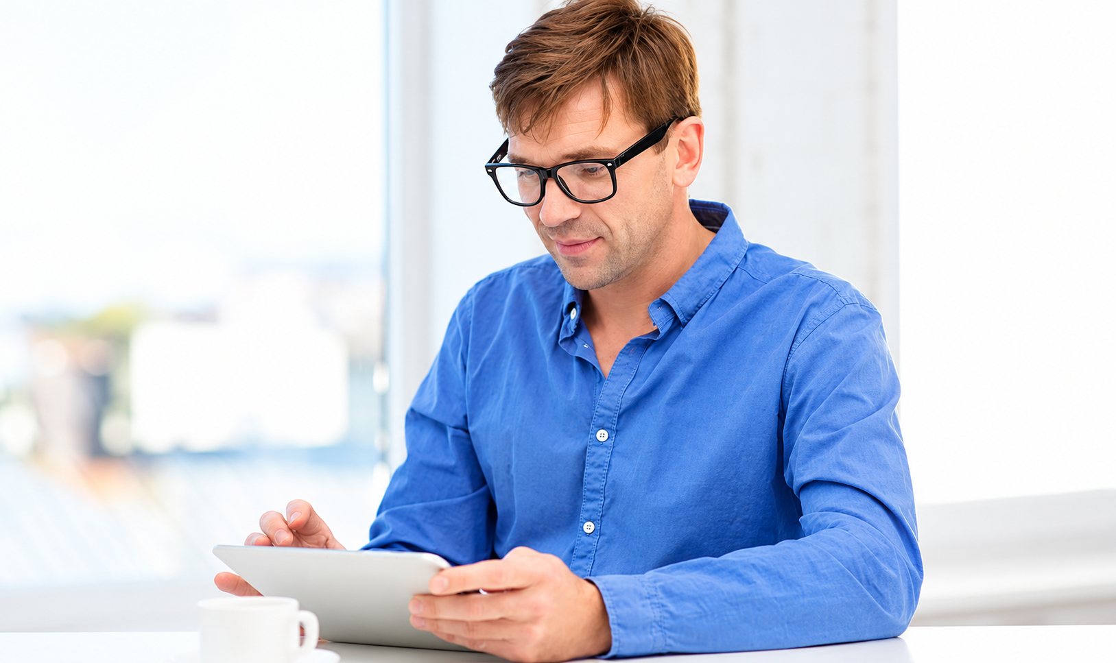 Un homme à lunettes noires et chemise bleue consulte une tablette assis à une table, près d'une grande fenêtre lumineuse.