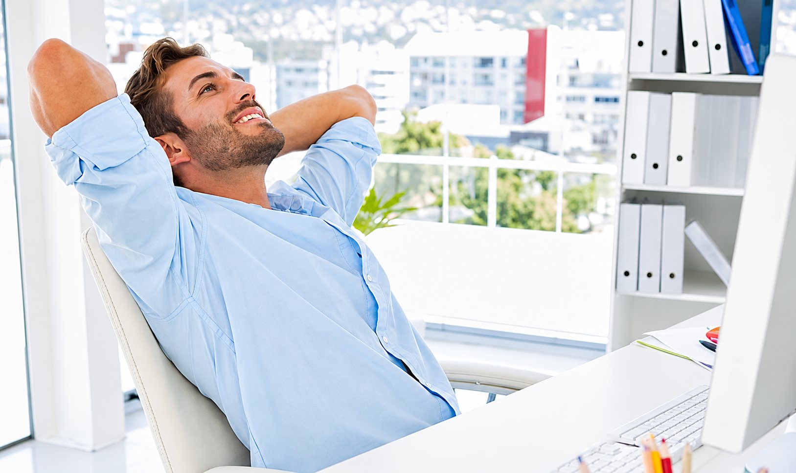 Un homme en chemise bleue claire sourit, adossé à sa chaise de bureau, les mains derrière la tête, dans un bureau lumineux avec des classeurs.