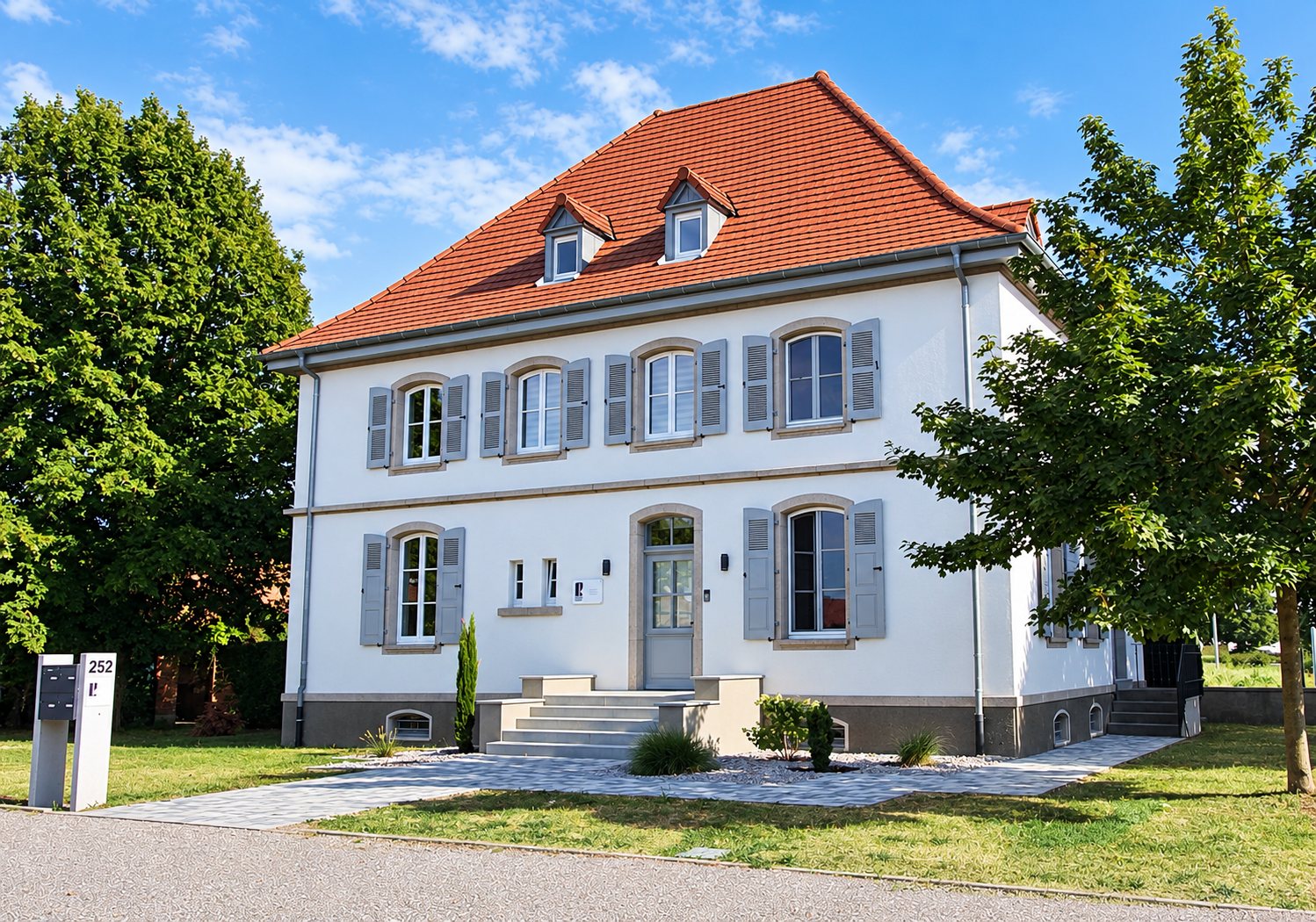 Façade d'une grande maison blanche rénovée avec volets gris, toit de tuiles rouges et lucarnes, entourée d'arbres et d'une pelouse.